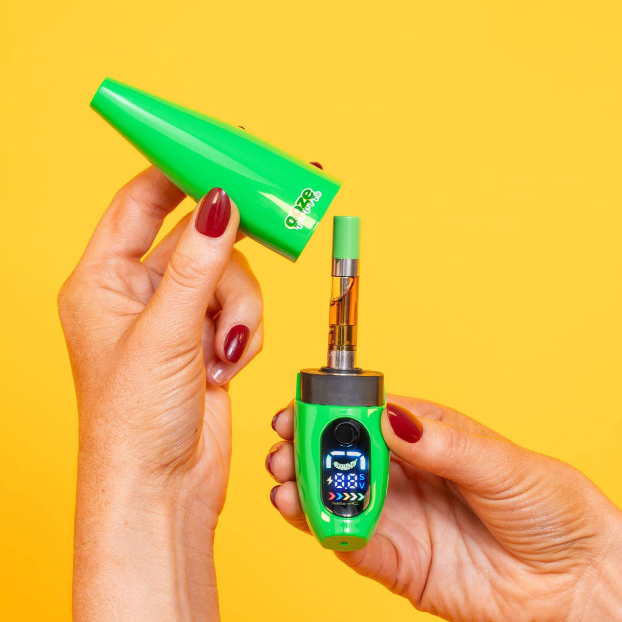 Green Ooze Peel banana vape device with digital display held by a hand and removing the cap to show the cartridge against a yellow background