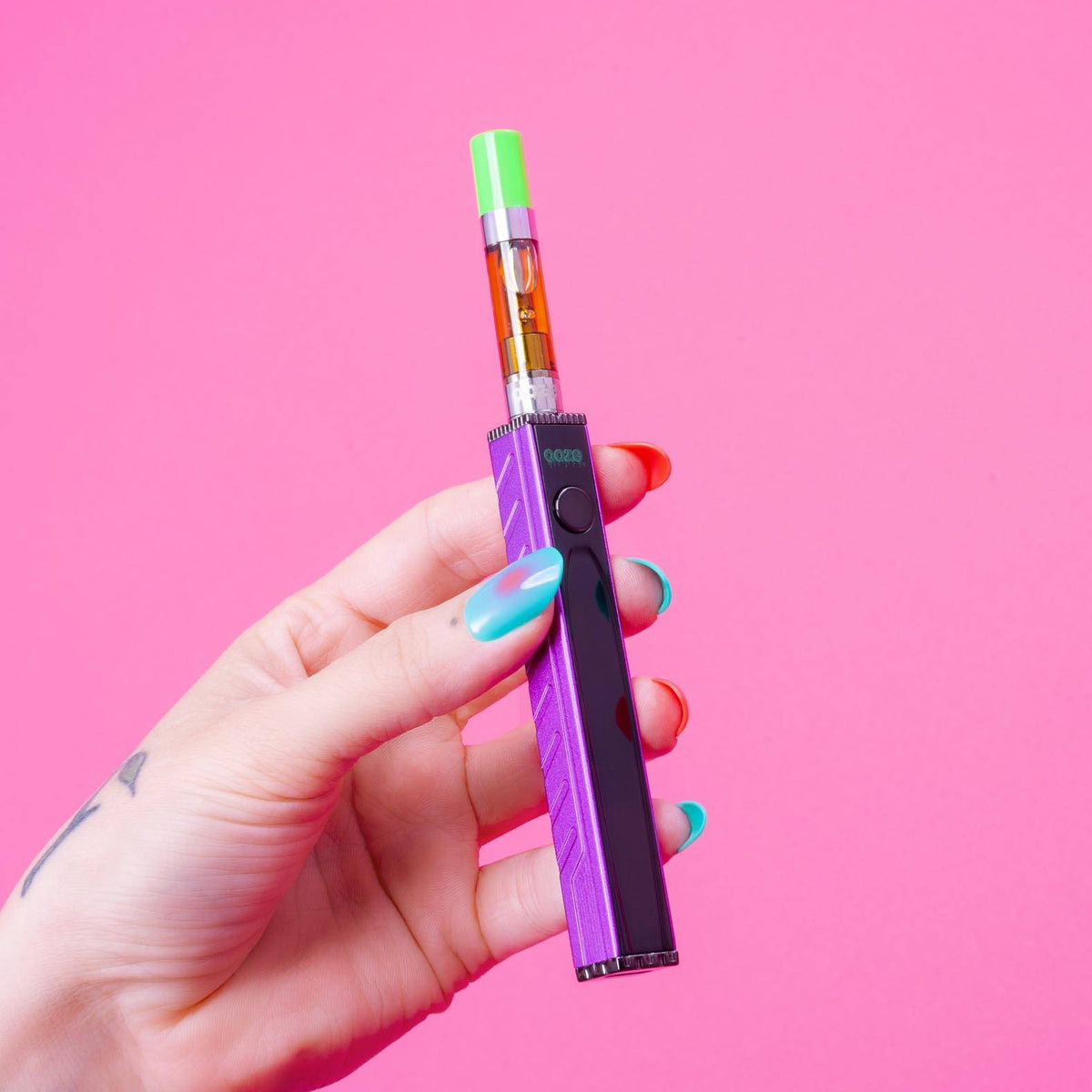A hand with colorful nail polish and a sun tattoo holds a square Ozoe Quad 2 purple and black vape device with a green mouthpiece against a solid pink background.