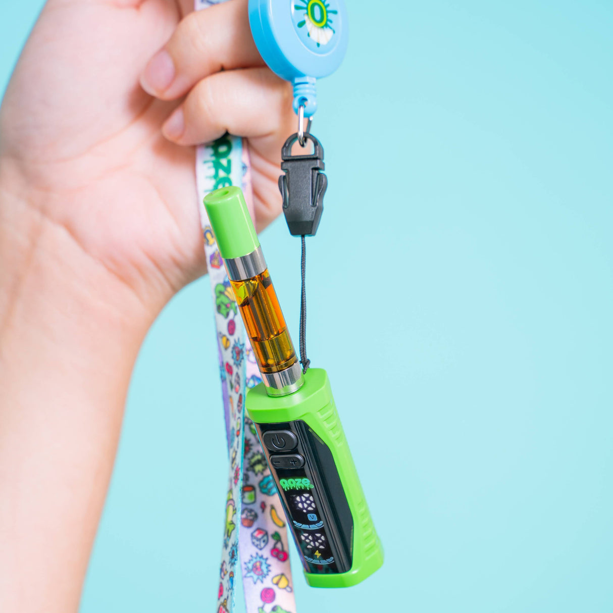 A hand holds a green Ooze Runt Mini vape device attached to a colorful patterned wrist strap with a black clip and blue retractable reel, against a light blue background.