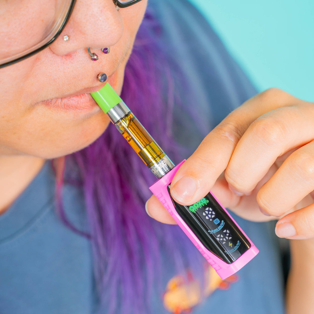 A close-up lifestyle shot of a person with purple hair and glasses vaping an oil cartridge. The Ooze Runt Mini vape device features a bright pink outer body, a black digital screen, and a green mouthpiece.