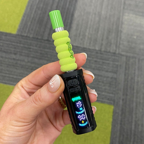 A hand holding a black Ooze Runt Mini Battery device with a bright lime green textured grip and a matching green mouthpiece, set against a gray and green carpeted floor.