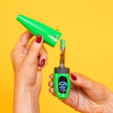 Green Ooze Peel banana vape device with digital display held by a hand and removing the cap to show the cartridge against a yellow background