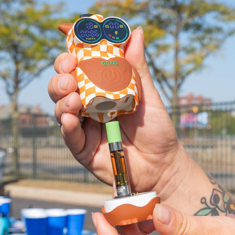 A hand holds an Ooze Hoot Character Vape device shaped like an owl, with orange and white checkered details, a brown owl face, and a 510 oil cartridge attached. The background is an outdoor blurred scene with trees and blue cups on the ground.