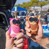 Two Ooze Hoot owl-shaped vape devices held in hands outdoors at a tailgate. One device is pink and cream, and the other is orange and white checkered.